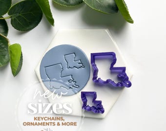 May include: Two purple plastic cookie cutters in the shape of the state of Louisiana. The cutters are on a white surface with a blue circle behind them. The text "new sizes, keychains, ornaments & more" is printed on the surface.