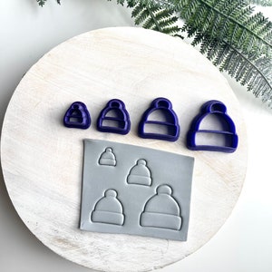 May include: A set of four blue plastic cookie cutters shaped like winter hats. The cutters are arranged in order of size, with the smallest cutter on the left and the largest cutter on the right. A sheet of grey polymer clay is shown with four impressions of the cookie cutters.