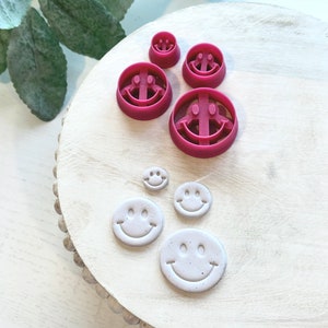 Könnte beinhalten: Ein Set mit fünf rosafarbenen Smiley-Ausstechern, mit drei größeren Ausstechern und zwei kleineren Ausstechern. Die Ausstecher befinden sich auf einer weißen Holzoberfläche mit einem perlenbesetzten Rand.