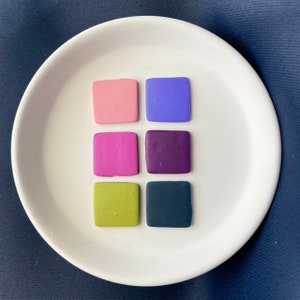 May include: Six small square tiles of different colors. The tiles are arranged in two rows of three. The colors are pink, purple, magenta, dark purple, green, and dark blue.