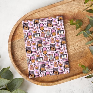May include: A pink notebook with a repeating pattern of Halloween-themed illustrations. The design includes pumpkins, ghosts, spiderwebs, and the word "spooky" in a decorative font. The notebook is on a wooden tray.
