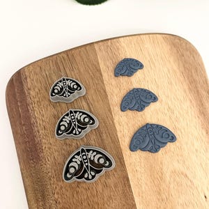 May include: Four butterfly-shaped cookie cutters, two in a light gray color and two in a dark blue color. The cutters are arranged in two rows on a wooden surface.