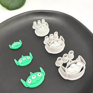 May include: Four cookie cutters in the shape of a three-eyed alien character from the movie Toy Story. The cutters are made of gray plastic and are arranged in a row on a black plate. The cutters are different sizes, with the largest cutter at the top of the image and the smallest cutter at the bottom. There are also three green alien cookie cutters on the plate.