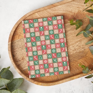 May include: A rectangular Christmas card with a festive pattern of red, green, and tan squares. Each square features a white outline of holiday icons like gingerbread men, snowflakes, and candy canes. The card is placed on a wooden tray.