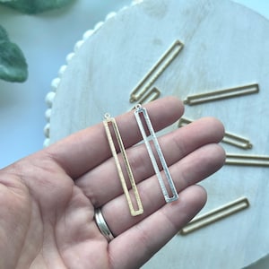 May include: Two metal rectangular pendant charms, one gold and one silver, with a textured surface. The charms have a loop at the top for attaching to jewelry.