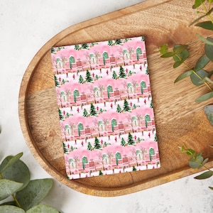 May include: Pink wrapping paper with a repeating pattern of a winter village scene. The pattern features small, colorful houses, trees, and people ice skating.