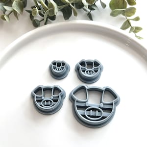 May include: Four gray pig-shaped cookie cutters of varying sizes are arranged on a white plate. The cutters have a simple design with a round face, ears, and a snout. Green leafy sprigs are in the background.