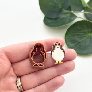 May include: A brown plastic Porg cookie cutter next to a Porg-shaped figurine. The figurine has a white body, brown and black head, and yellow feet. The cookie cutter is in the shape of the Porg. The background is white with green leaves.