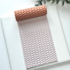 May include: A light pink silicone rolling pin with a diamond pattern. The rolling pin is on a white surface.
