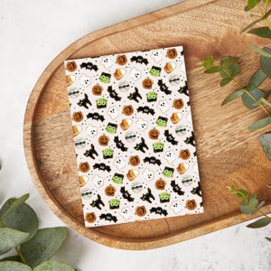 May include: A notebook featuring a repeating pattern of Halloween illustrations. The design includes pumpkins, bats, ghosts, Frankenstein's monster, and candy corn in orange, black, green, and white on a light beige background. The notebook is on a wooden tray.