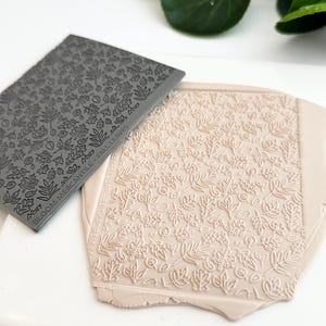 May include: A gray silicone stamp with a pattern of leaves and flowers. The stamp is being used to create an impression in a light pink clay surface. The text "BLOOM MERCANTILE FALL LEAVES" is visible on the stamp.