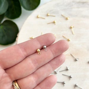 May include: Close-up of gold and silver-colored ball-end earring posts with small loops. Several posts are scattered on a light-colored wooden surface. The earrings are small and delicate, suitable for jewelry making or crafting.