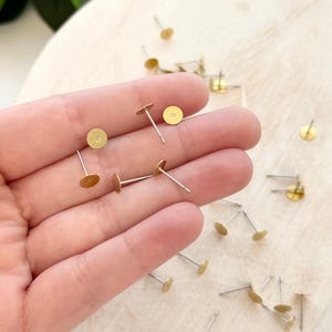 May include: A collection of gold-colored earring posts with flat, round tops and silver-colored posts. The earring findings are scattered on a light-colored wooden surface. Some of the earring posts are held in a hand.