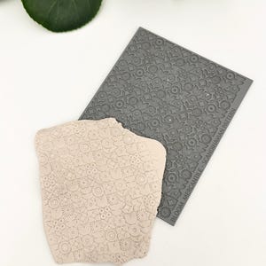 May include: A gray silicone stamp with a repeating pattern of small flowers and circles. The text "BLOOM MERCANTILE - SCANDINAVIAN PATTERN" is printed on the stamp. A light pink clay impression of the stamp is shown next to it.