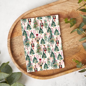 May include: A white card with a repeating pattern of watercolour nutcracker soldiers and Christmas trees. The nutcrackers are wearing red and gold uniforms and the trees are green with pink accents.