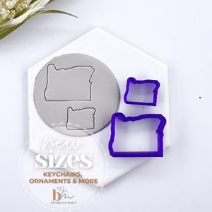 Puede incluir: Tres cortadores de galletas de plástico morado en forma del estado de Oregón. Los cortadores están sobre una superficie blanca con el texto "new sizes, keychains, ornaments & more" impreso debajo.