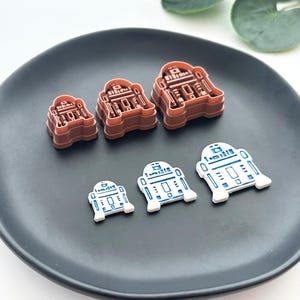 Puede incluir: Una colección de cortadores de galletas y decoraciones de fondant con forma de R2-D2, marrones y blancos. Los cortadores de galletas son marrones y las decoraciones de fondant son blancas con detalles azules. Los artículos están colocados en un plato gris oscuro.