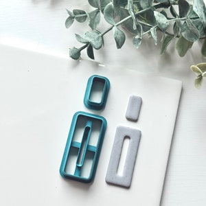 May include: Two cookie cutters, one blue and one gray, shaped like rectangles with a rectangular hole in the center. The blue cutter has a smaller rectangular hole inside the larger one.