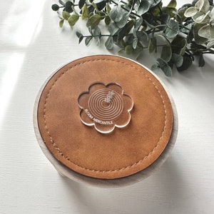 Op de afbeelding: Een ronde, houten onderzetter met een gestikte, bruine leren bovenkant. Een helder, bloemvormig acryl stuk met concentrische cirkels en de tekst "BLOOM MERCANTILE" is gecentreerd op het leer. Groen is op de achtergrond.