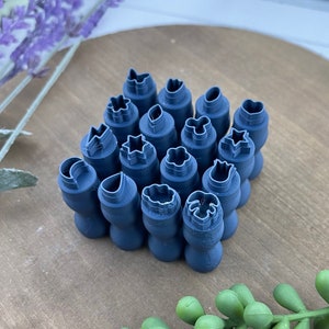 May include: A set of 18 blue polymer clay stamps with various shapes, including stars, flowers, leaves, and a crescent moon. The stamps are arranged in a circular pattern on a wooden surface.