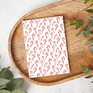 May include: A rectangular card with a repeating pattern of red and white candy canes on a white background. The card is placed on a wooden tray with a light-colored surface and green foliage.