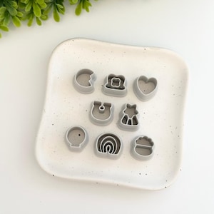 May include: A set of nine gray plastic cookie cutters in various shapes, including a heart, a rainbow, a mushroom, a star, a balloon, a bear, a flower, a hat, and a cupcake. The cutters are arranged on a white speckled plate.