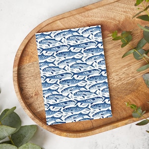Peut inclure: Un carnet rectangulaire avec un motif répétitif de poissons bleus sur fond blanc. Les poissons sont stylisés avec des écailles et des nageoires détaillées. Le carnet est posé sur un plateau en bois, avec des feuilles vertes en arrière-plan.