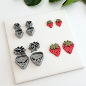 May include: A set of four gray polymer clay cutters shaped like strawberries, with two cutters featuring a leaf design. Two red polymer clay strawberries with green leaves are also shown.