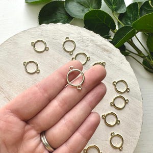 May include: Gold-coloured metal connector rings with small loops on each side. The rings are arranged on a light-coloured wooden surface, with green leafy plants in the background. The rings are used for jewellery making.