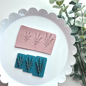May include: Three blue polymer clay stamps with a design of lavender sprigs. The stamps are on a white plate with a scalloped edge.