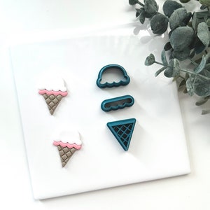 Può includere: Un set di quattro tagliabiscotti in plastica blu-verde a forma di coni e palline di gelato. I tagliabiscotti sono su una superficie bianca.