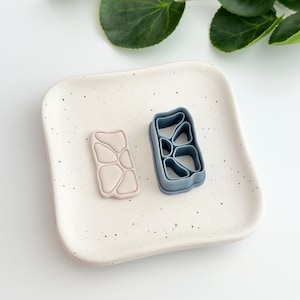 May include: Two silicone cookie cutters on a white speckled plate. The cutter on the left is pink and has a textured design. The cutter on the right is blue and has a geometric design.