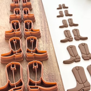 May include: A set of brown plastic cowboy boot cookie cutters on a wooden surface. The cutters are arranged in rows, with the largest cutters at the bottom and the smallest cutters at the top. There are also several brown sugar cookie boots on a white surface.