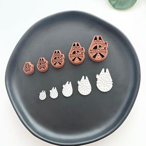 May include: A collection of brown and white Millennium Falcon-shaped cookie cutters in various sizes, arranged on a dark gray plate. The cutters are made of plastic and are designed for baking themed cookies. The arrangement shows the cutters in ascending order of size.