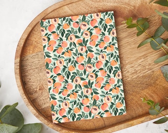 May include: A rectangular notebook with a peach and leaf pattern. The design features numerous peaches in shades of orange and pink, interspersed with green leaves, set against a light background. The notebook is placed on a wooden tray.