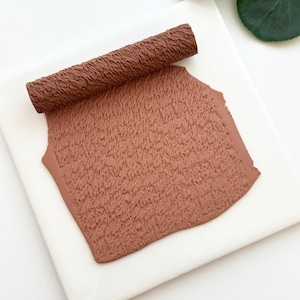 May include: A textured, brown clay roller and a sheet of clay sit on a white surface. The clay has a woven texture, and the roller is cylindrical. The clay and roller are a warm, earthy brown color.