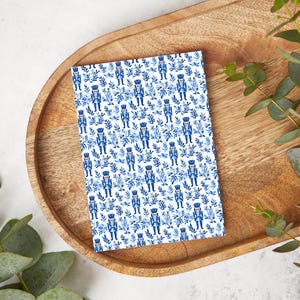 May include: A rectangular card with a blue and white pattern of toy soldiers and foliage. The card is resting on a wooden tray, with green leaves in the background.