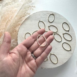 May include: A collection of gold-colored oval pendant blanks. One is held in a hand, with others scattered on a light-colored wooden surface. The pendants have a small loop at the top for attaching a chain or cord. The background includes dried ornamental grass.
