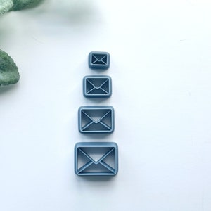 May include: Four blue envelope-shaped cookie cutters in varying sizes. The cutters are square with a detailed envelope design in the center. The cutters are arranged vertically on a white surface.