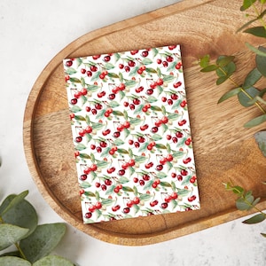 May include: A rectangular card with a repeating pattern of red cherries and green leaves on a white background. The card is placed on a wooden tray with a light brown finish. Green foliage is visible in the background.