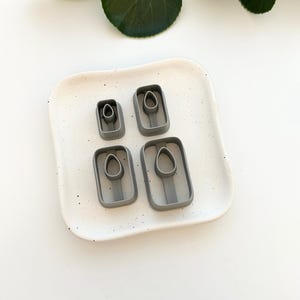 May include: Four gray plastic cookie cutters shaped like an oval with a smaller oval inside. The cutters are arranged in two rows of two on a white speckled surface.