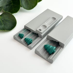 May include: A gray metal case with a sliding lid, containing three small green tree-shaped earplugs. The case has a textured surface and a small opening on the top.