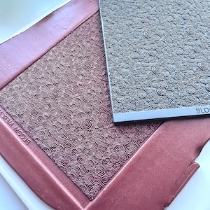 Puede incluir: Primer plano de dos alfombras de relieve de silicona con un patrón repetitivo de círculos concéntricos. La alfombra superior es gris y la inferior es de color burdeos oscuro. El texto "BLOOM WHERE YOU ARE PLANTED" es visible en la alfombra inferior.