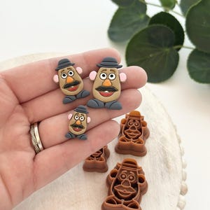 Può includere: Una collezione di ciondoli e tagliabiscotti in argilla polimerica a tema Mr. Potato Head. I ciondoli hanno la forma del personaggio con cappello nero, baffi e guance rosee. I tagliabiscotti sono marroni e raffigurano il volto del personaggio.