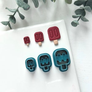 May include: An overhead shot shows three sizes of popsicle-shaped cookie cutters in teal and three red popsicle-shaped objects. The red popsicles have a textured surface and tan sticks. The teal cutters are plastic and have a popsicle shape.