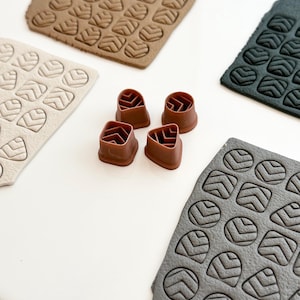 May include: Four brown plastic stamps with a chevron pattern, used to create impressions in clay. The stamps are arranged in a square shape on a white surface. Three sheets of clay are visible in the background, each with a different color and a chevron pattern.
