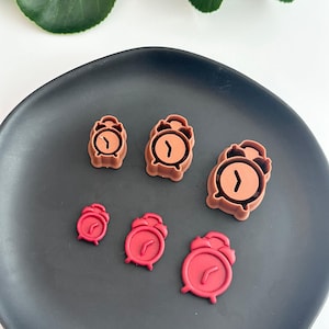 May include: Five different sized cookie cutters in the shape of an alarm clock. The cutters are brown and red. The cutters are on a black plate.