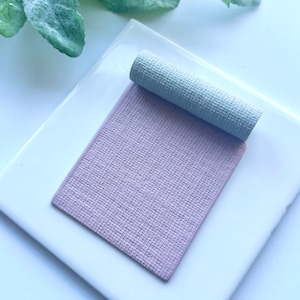 May include: A close-up of a light purple textured fabric sample. The fabric is rolled up and placed on a white surface.