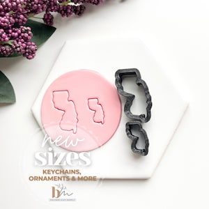 May include: Two black cookie cutters in the shape of the state of New Jersey. The cutters are on a white surface with a pink circle in the center. The circle has two outlines of the state of New Jersey in pink. The text "new sizes" is written in black below the circle. The text "keychains, ornaments & more" is written in black below the text "new sizes". The text "bh" is written in black below the text "keychains, ornaments & more".
