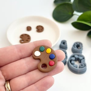 Puede incluir: Una pequeña paleta de artista marrón con puntos de pintura coloridos en rojo, azul, amarillo, verde y blanco. Varias paletas y cortadores grises más están en el fondo. La paleta se sostiene en una mano.